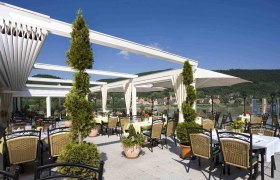 Sonnige Terrasse mit Tischen und Stühlen, Blick auf Fluss und Hügel., © Hotel Donauterrasse