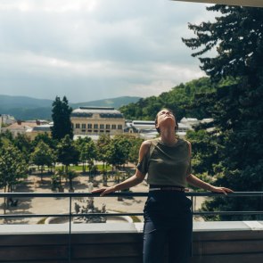 Eine Frau steht am Balkon eines Hotels. Im Hintergrund sieht man das Casino Baden und den Kurpark.