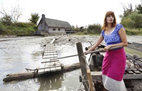 Frau in traditioneller Kleidung auf einem Boot vor einem Holzhaus am Flussufer., © Donau Niederösterreich, Steve Haider