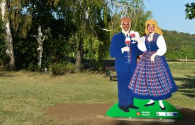 Pappfiguren in Tracht mit Weingläsern, Kreuz im Hintergrund, sonniger Park., © Donau Niederösterreich, Steve Haider