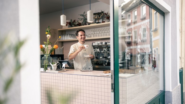 Feinbäcker Matthias Krenn steht mit einer Tasse Kaffee in seiner Bäckerei und lächelt in Richtung Schaufenster nach draußen